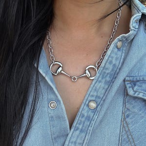 May include: Silver-toned necklace featuring a horse bit design. The necklace has a chain link style and a central bit element with two circular rings and a connecting bar. The jewelry is worn against a light blue denim shirt.