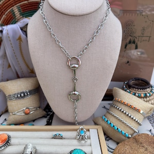 May include: A silver-toned necklace with a horse bit pendant is displayed on a mannequin. The jewelry display includes several bracelets and rings with turquoise and coral stones. A wooden tray holds the rings.