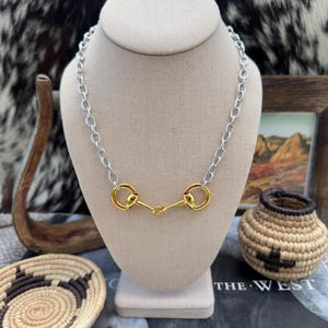 May include: A silver chain necklace with a gold-colored horse bit pendant. The necklace is displayed on a beige bust, with a rustic theme. The background includes a small woven basket, a framed landscape, and a cowhide pattern.