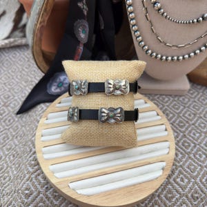 May include: Two black leather bracelets with silver-toned bow-shaped clasps, each featuring a small turquoise stone. The bracelets rest on a burlap pillow, displayed on a wooden tray with white inserts. A black patterned scarf and silver necklaces are in the background.