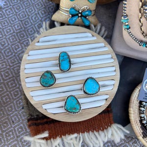 May include: A display of turquoise jewelry, including rings and butterfly-shaped bracelets. The rings feature silver settings and various turquoise stone shapes. The bracelets have turquoise butterfly designs. The jewelry is displayed on a wooden stand.