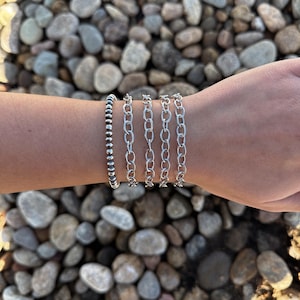 May include: Five silver bracelets stacked on a person's wrist. The bracelets are all different styles, including a beaded bracelet and several chain bracelets.