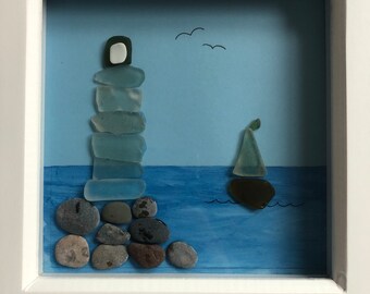 May include: A framed artwork featuring a lighthouse constructed from blue glass pieces resting on a bed of grey and brown stones. A small sailboat crafted from blue glass is sailing on the blue water. The background is a light blue sky with two black birds in flight.