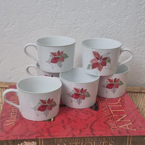 May include: A set of six white ceramic mugs with a poinsettia design in red and green. The mugs are stacked on a red book titled "The Complete Guide to Antiques". The mugs have a simple, elegant design, perfect for holiday gatherings.