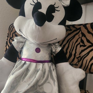 May include: A large plush toy of Minnie Mouse wearing a silver dress with white polka dots and a purple sash. She has a black bow with silver accents in her hair.