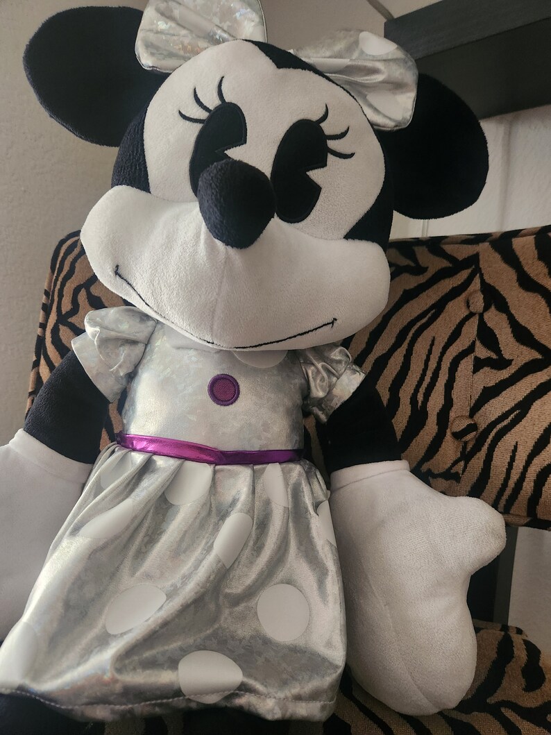 May include: A large plush toy of Minnie Mouse wearing a silver dress with white polka dots and a purple sash. She has a black bow with silver accents in her hair.