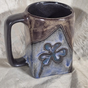 May include: A handmade ceramic mug with a square shape and a dark handle. The mug features a blue and gray floral design on a blue and brown background. The mug is approximately 4 inches tall.