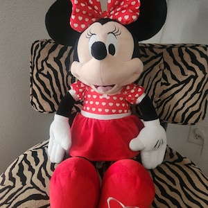 May include: A large plush Minnie Mouse toy wearing a red dress with white hearts, a red bow with white hearts, and red shoes with white hearts.