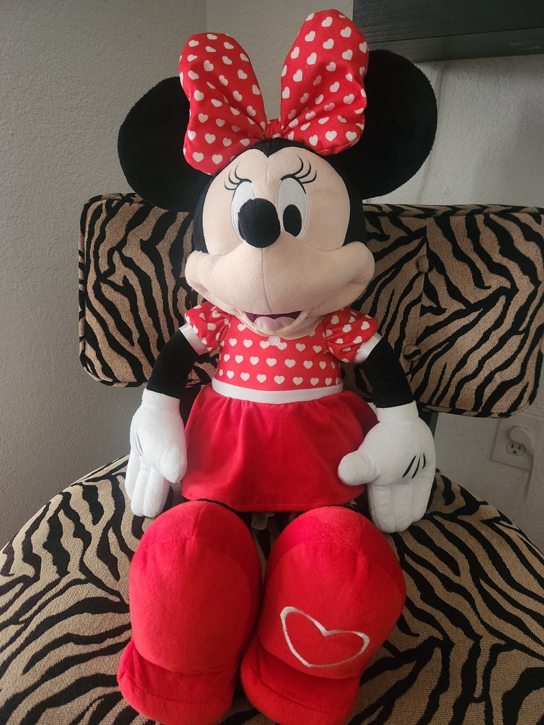 May include: A large plush Minnie Mouse toy wearing a red dress with white hearts, a red bow with white hearts, and red shoes with white hearts.
