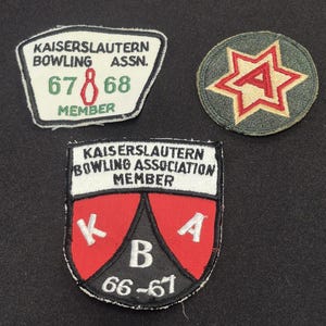May include: Three embroidered fabric patches. One is a white trapezoid with "KAISERSLAUTERN BOWLING ASSN. 67 68 MEMBER" and a red bowling pin. One is a circular olive green patch with a red and tan star and letter A. One is a shield with "KAISERSLAUTERN BOWLING ASSOCIATION MEMBER" and "K A B 66-67".