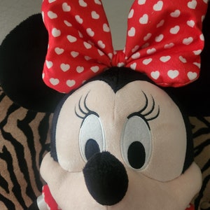 May include: A plush toy of a cartoon mouse with large white eyes, a black nose, and a red bow with white polka dots. The mouse is wearing a red dress with white polka dots.