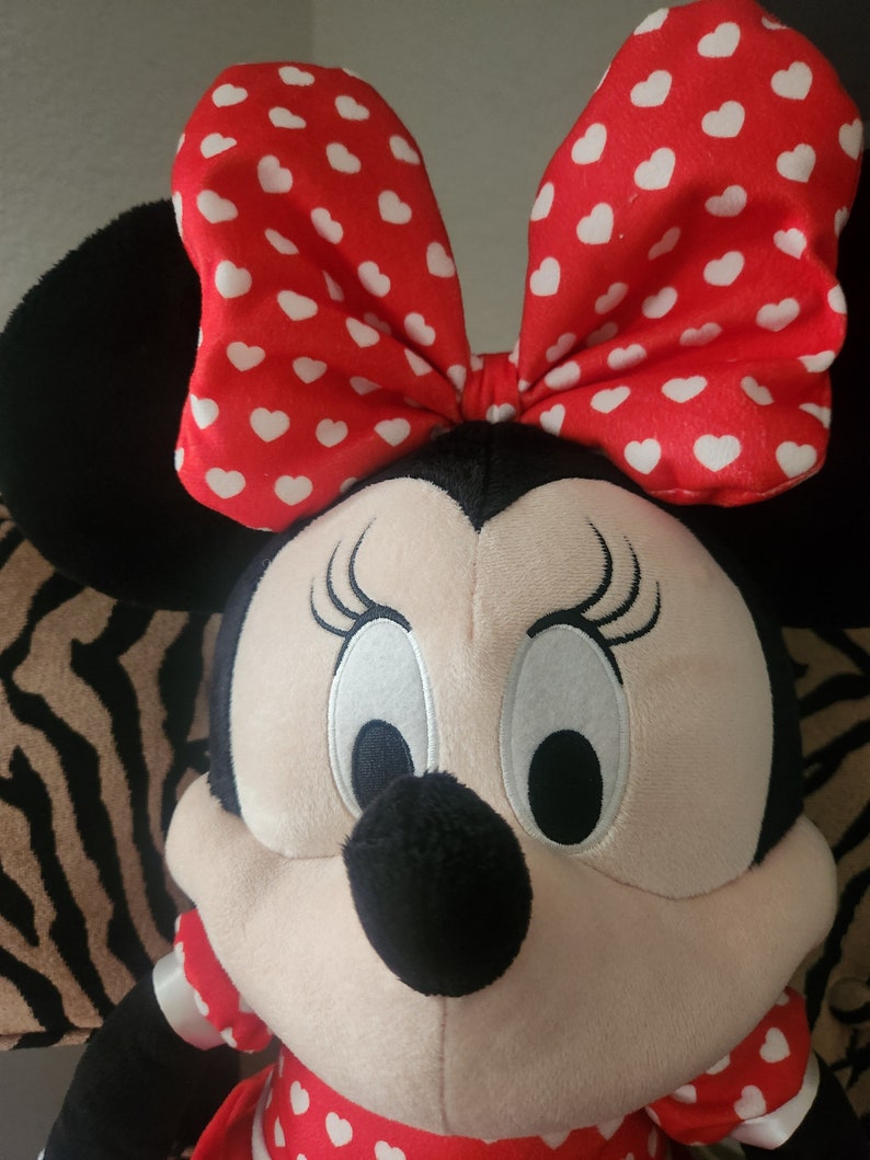 May include: A plush toy of a cartoon mouse with large white eyes, a black nose, and a red bow with white polka dots. The mouse is wearing a red dress with white polka dots.