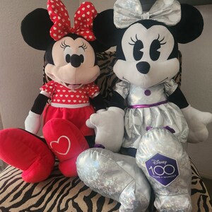 May include: Two plush Minnie Mouse toys. One is wearing a red dress with white polka dots and a red bow. The other is wearing a silver dress with a silver bow and a purple patch that says "Disney 100 Years of Wonder".