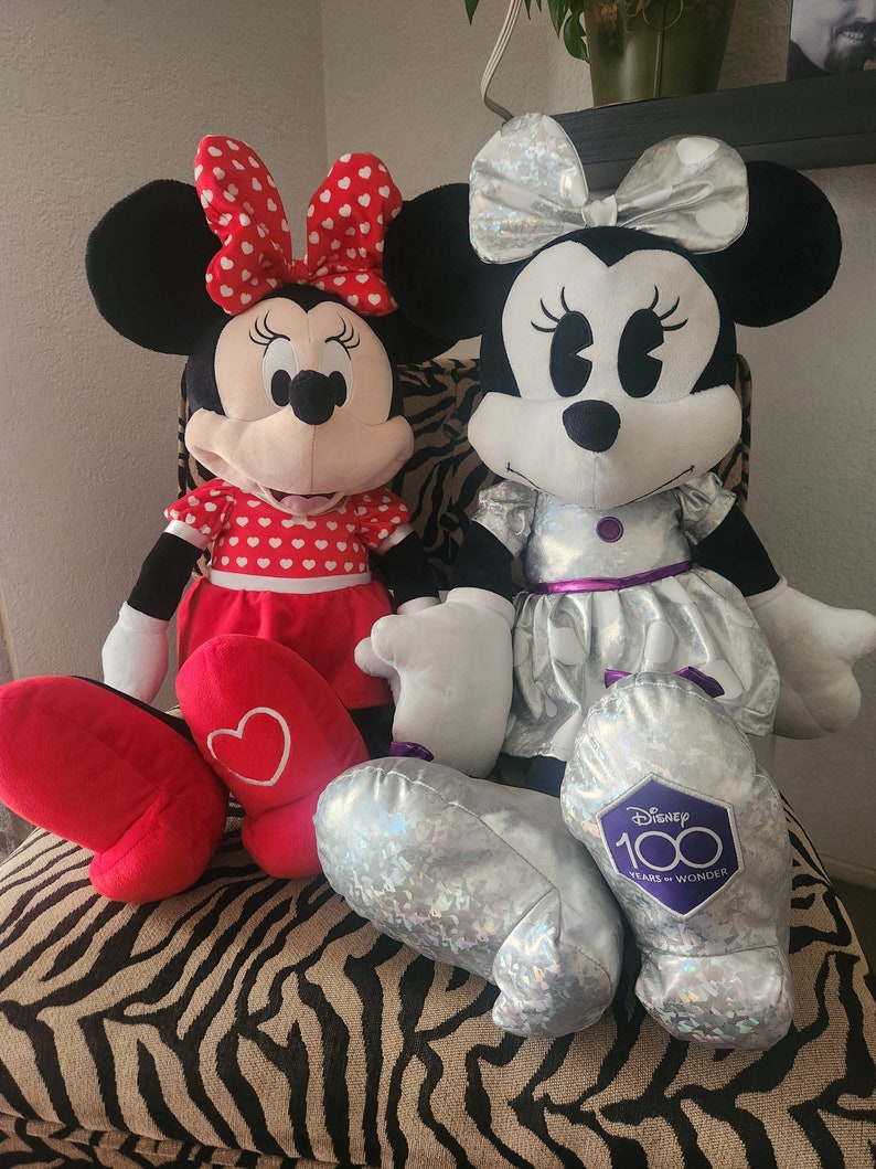 May include: Two plush Minnie Mouse toys. One is wearing a red dress with white polka dots and a red bow. The other is wearing a silver dress with a silver bow and a purple patch that says "Disney 100 Years of Wonder".