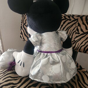 May include: A plush Minnie Mouse toy wearing a silver dress with white polka dots and a purple sash. The toy has black ears and a black tail.