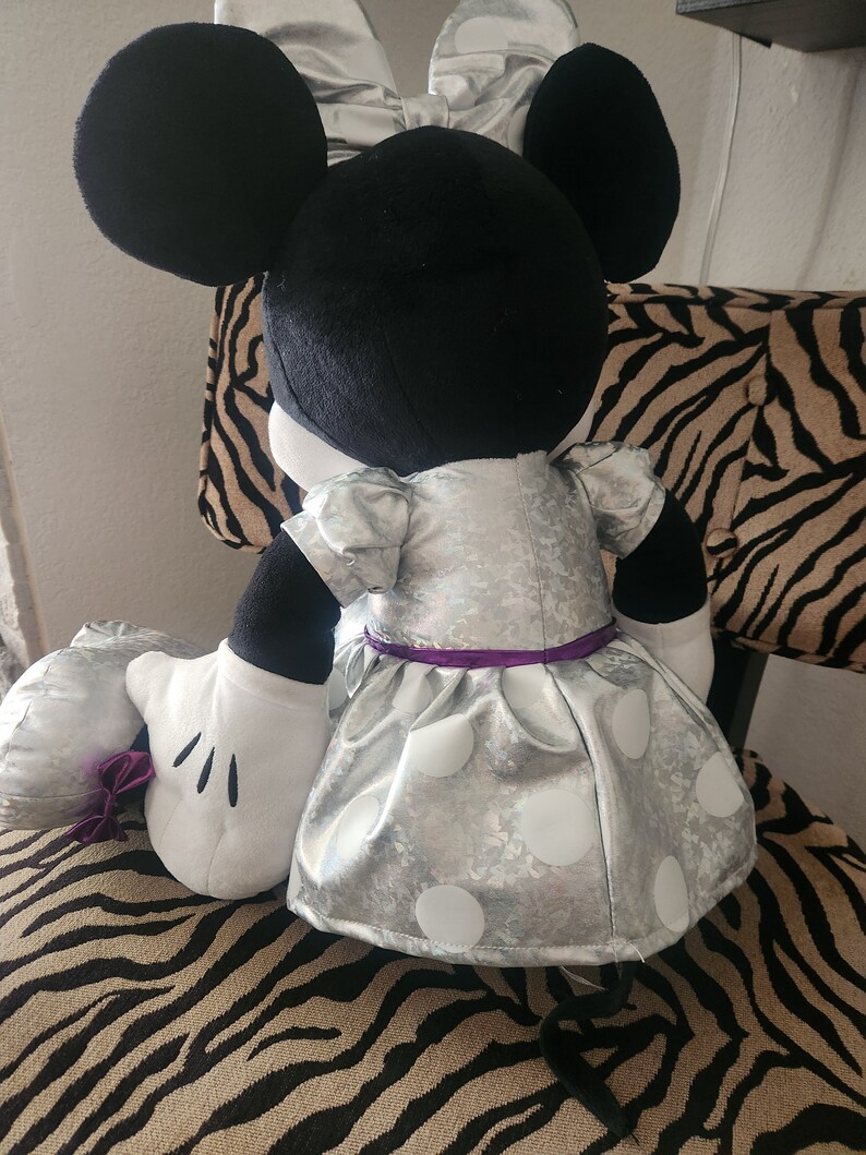 May include: A plush Minnie Mouse toy wearing a silver dress with white polka dots and a purple sash. The toy has black ears and a black tail.