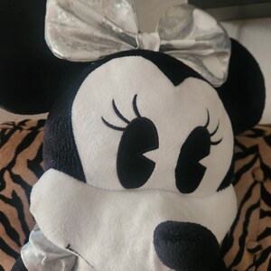 May include: A large plush toy of a cartoon mouse with black ears and a silver bow. The mouse has white fur and black eyes with long eyelashes. The mouse is sitting on a striped surface.