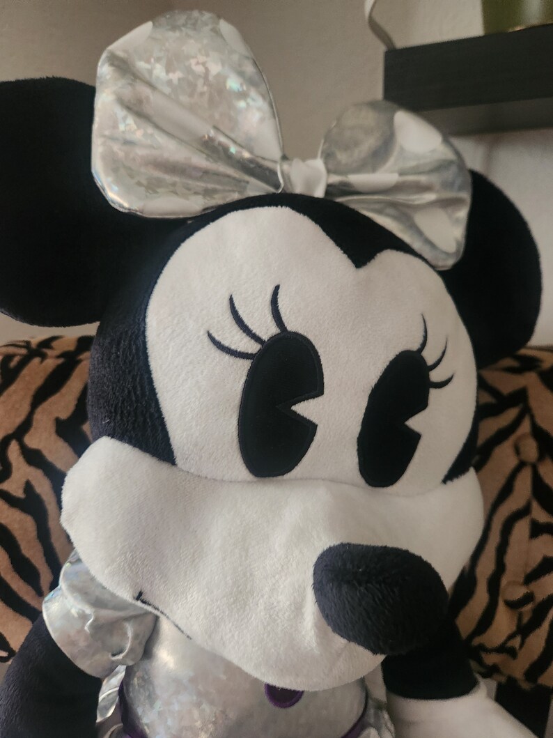 May include: A large plush toy of a cartoon mouse with black ears and a silver bow. The mouse has white fur and black eyes with long eyelashes. The mouse is sitting on a striped surface.