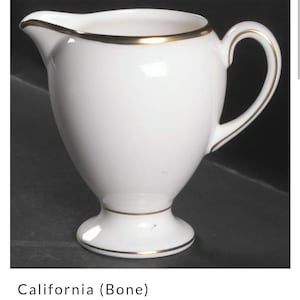 May include: A white globe-shaped creamer with a gold trim around the top and base. The creamer has a handle and is made by Wedgwood.