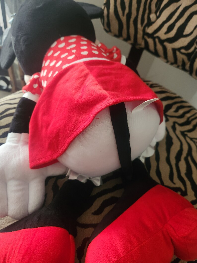 May include: A large plush toy of a cartoon mouse wearing a red dress with white polka dots. The mouse is sitting on a zebra-print fabric.