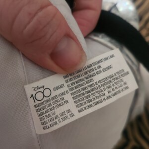 May include: A white tag with black text that reads "Disney 100" and includes care instructions and manufacturing information. The tag is attached to a plush toy.