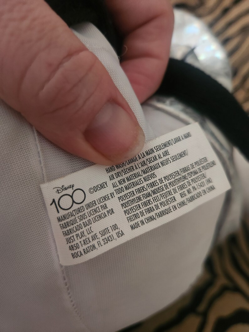 May include: A white tag with black text that reads "Disney 100" and includes care instructions and manufacturing information. The tag is attached to a plush toy.