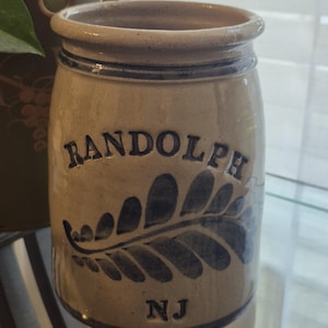 May include: A tan stoneware jar with a dark blue fern design and the word "RANDOLPH" arched across the front. The letters "NJ" are below the fern. The jar has a dark blue rim and a slightly flared top.