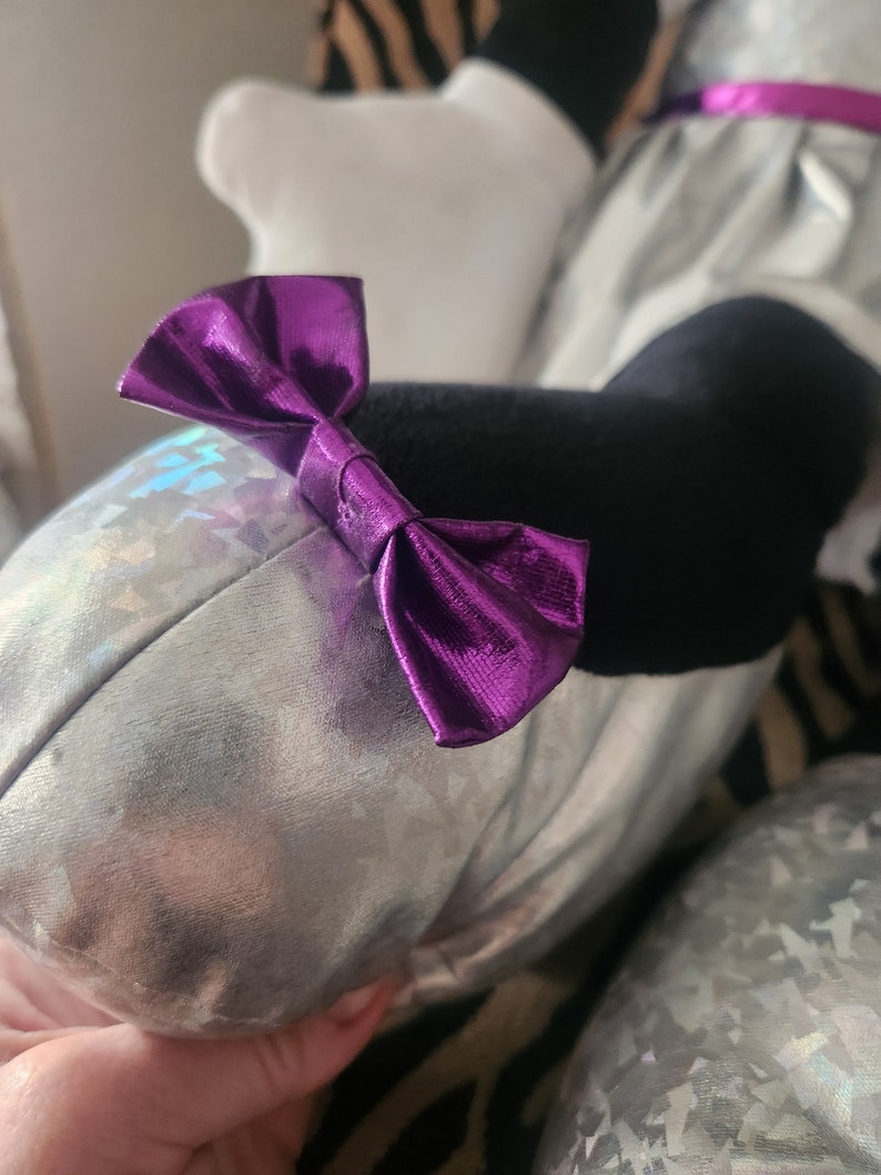May include: A silver plush toy with a purple bow. The toy has a holographic iridescent finish.
