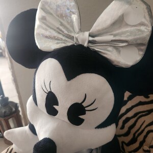May include: A plush black and white Minnie Mouse toy with a silver bow with white polka dots. The toy is sitting on a zebra-print surface.