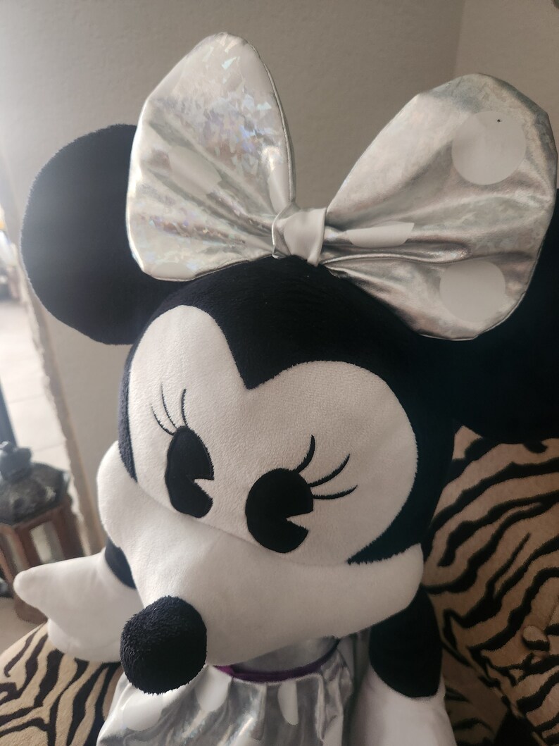 May include: A plush black and white Minnie Mouse toy with a silver bow with white polka dots. The toy is sitting on a zebra-print surface.