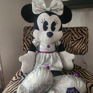 May include: A large plush Minnie Mouse doll wearing a silver sequined dress with a purple sash and a silver bow. The doll is sitting on a zebra-print chair and has a silver and purple 100th anniversary Disney logo on one foot.