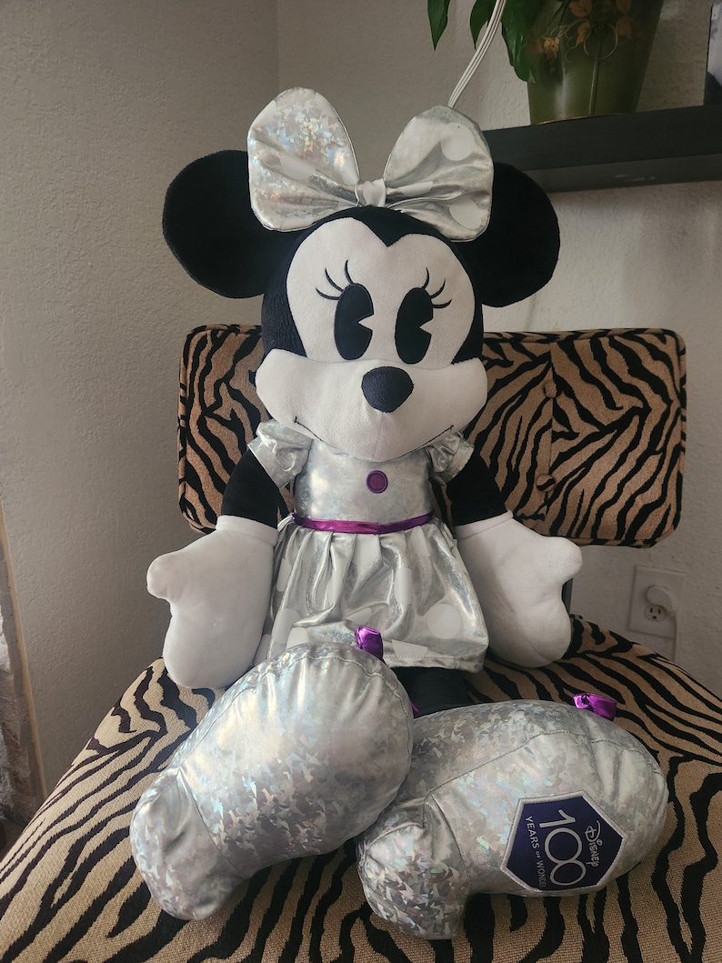 May include: A large plush Minnie Mouse doll wearing a silver sequined dress with a purple sash and a silver bow. The doll is sitting on a zebra-print chair and has a silver and purple 100th anniversary Disney logo on one foot.
