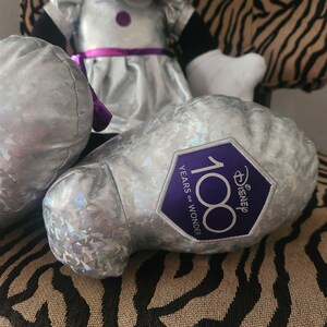 May include: A silver plush toy with a purple bow and a silver and purple patch that says "Disney 100 Years of Wonder".