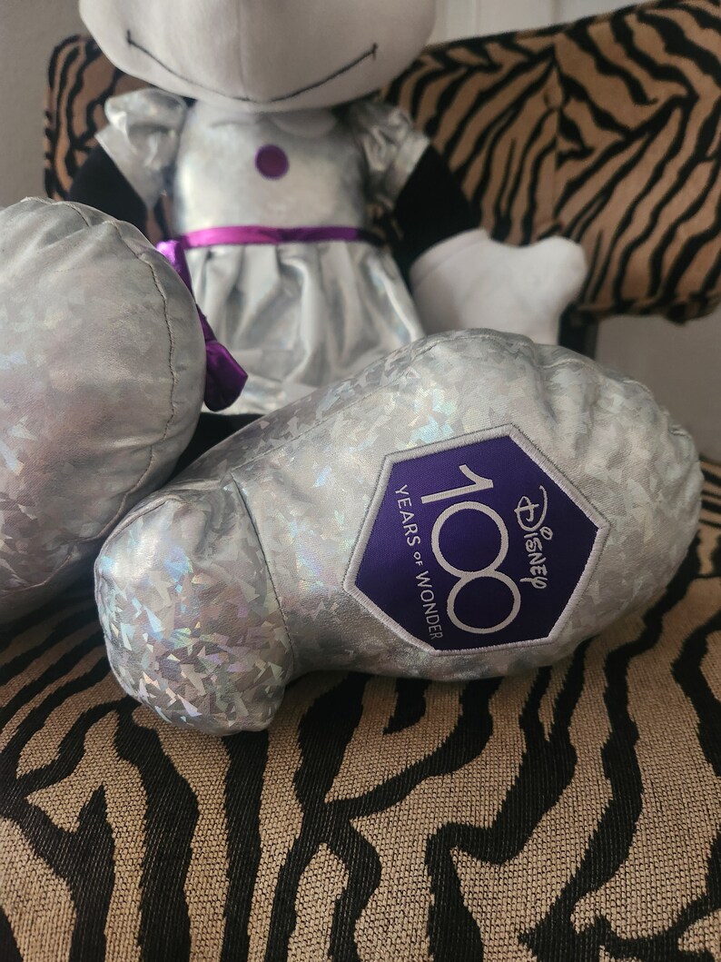 May include: A silver plush toy with a purple bow and a silver and purple patch that says "Disney 100 Years of Wonder".
