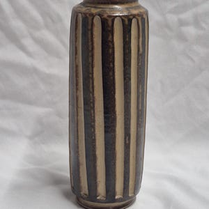 May include: A tall, cylindrical ceramic vase featuring a repeating pattern of vertical, recessed grooves. The surface displays a two-tone color scheme with alternating dark brown and light beige stripes, finished with a smooth, glazed texture.