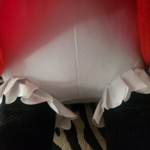 May include: A plush toy with a red and white dress and white ruffled trim. The toy is sitting on a striped fabric.