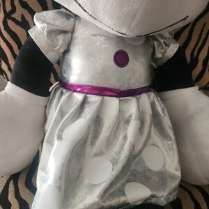 May include: A plush toy with a silver dress with white polka dots and a purple ribbon. The dress has a purple button on the front.