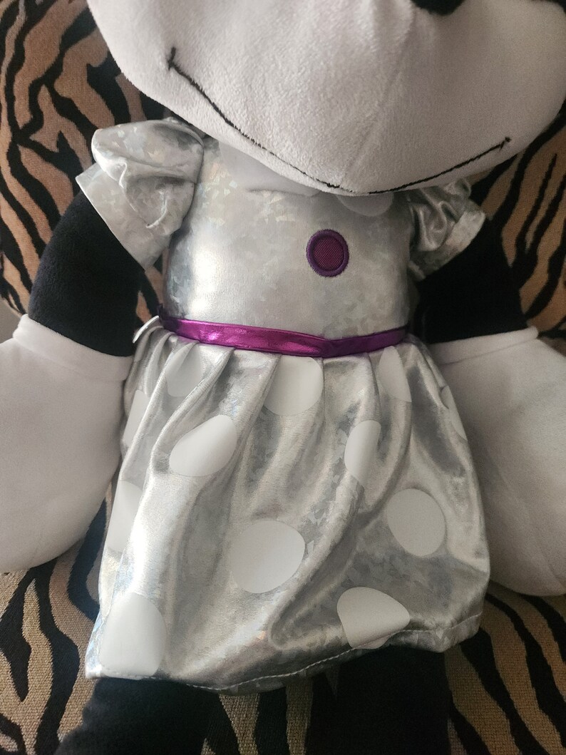 May include: A plush toy with a silver dress with white polka dots and a purple ribbon. The dress has a purple button on the front.