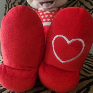 May include: Red plush slippers with a white heart embroidered on one slipper. The slippers are sitting on a zebra-print surface.