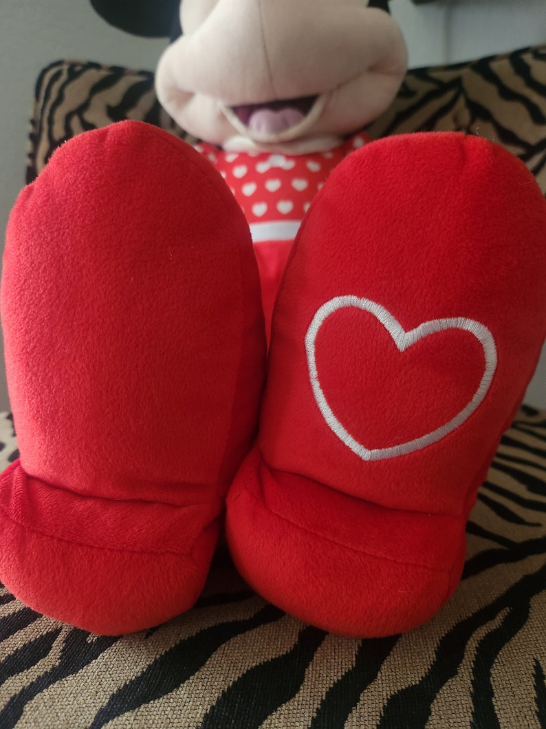 May include: Red plush slippers with a white heart embroidered on one slipper. The slippers are sitting on a zebra-print surface.