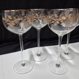 May include: A set of four clear glass stemmed goblets featuring a delicate gold floral vine pattern encircling the upper bowl and a thin gold rim. Each glass has a slender stem and a circular base, designed for serving beverages with elegance.