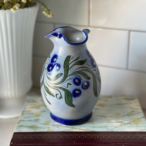 May include: A ceramic vase with a light blue speckled finish, adorned with blue floral and green leaf designs. The vase has a ruffled rim with a dark blue trim and a matching base. A white vase and flowers are in the background.