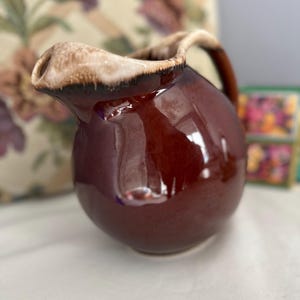 May include: A glossy, brown ceramic jug with a rounded body and a handle. The rim and spout have a lighter, tan-coloured glaze. The jug is likely for serving beverages and has a classic, vintage aesthetic.