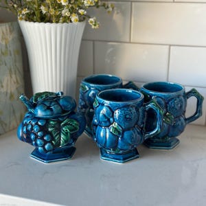 May include: A collection of four blue ceramic mugs and a matching creamer, each adorned with a grape and leaf motif. The mugs feature handles and sit on a hexagonal base. The creamer has a lid and spout. A white vase with flowers is in the background.
