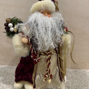 May include: A festive Santa Claus ornament. The figure is dressed in a burgundy velvet coat with white fur trim, a cream-colored embroidered robe, and a white fur hat. He holds a staff and a small bouquet of berries.