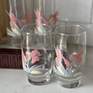 May include: Four clear glass tumblers with a floral design. Each glass has a pink iris flower with grey and yellow accents. The glasses are cylindrical with a flared base, ideal for serving drinks. The height is approximately 15 cm.