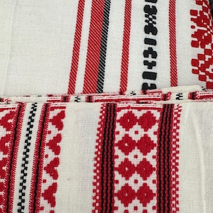 May include: White fabric with red and black embroidered stripes and geometric patterns. The design features vertical stripes of varying widths, with detailed red diamond and floral motifs. The fabric appears to be woven, with a textured surface.