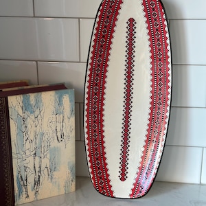 May include: An oval serving platter with a black edge and a white interior. The platter is decorated with a red and black geometric pattern, resembling folk art. The design runs along the sides and down the centre. The platter is displayed on a white surface.