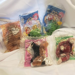 May include: Five small plush toys in individual clear plastic bags with colorful packaging. The toys are various colors and feature different characters. The bags have warnings and instructions in English and Spanish.
