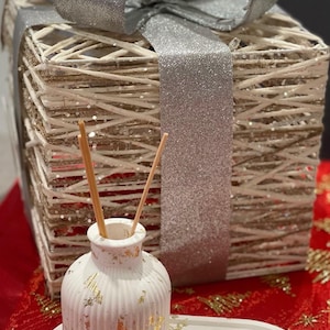 Può includere: Un diffusore in ceramica bianca con dettagli dorati si trova su un vassoio ovale bianco. Il diffusore ha due bastoncini di legno al suo interno. Il vassoio è su una superficie rossa con glitter dorati. Una scatola regalo in vimini bianca con un nastro argentato si trova sullo sfondo.